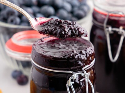 Spoon lifting a thick, glossy scoop of homemade Blueberry Butter above a clip-top glass jar, showcasing the rich texture of the slow cooker fruit spread with fresh blueberries and filled jars in the background.