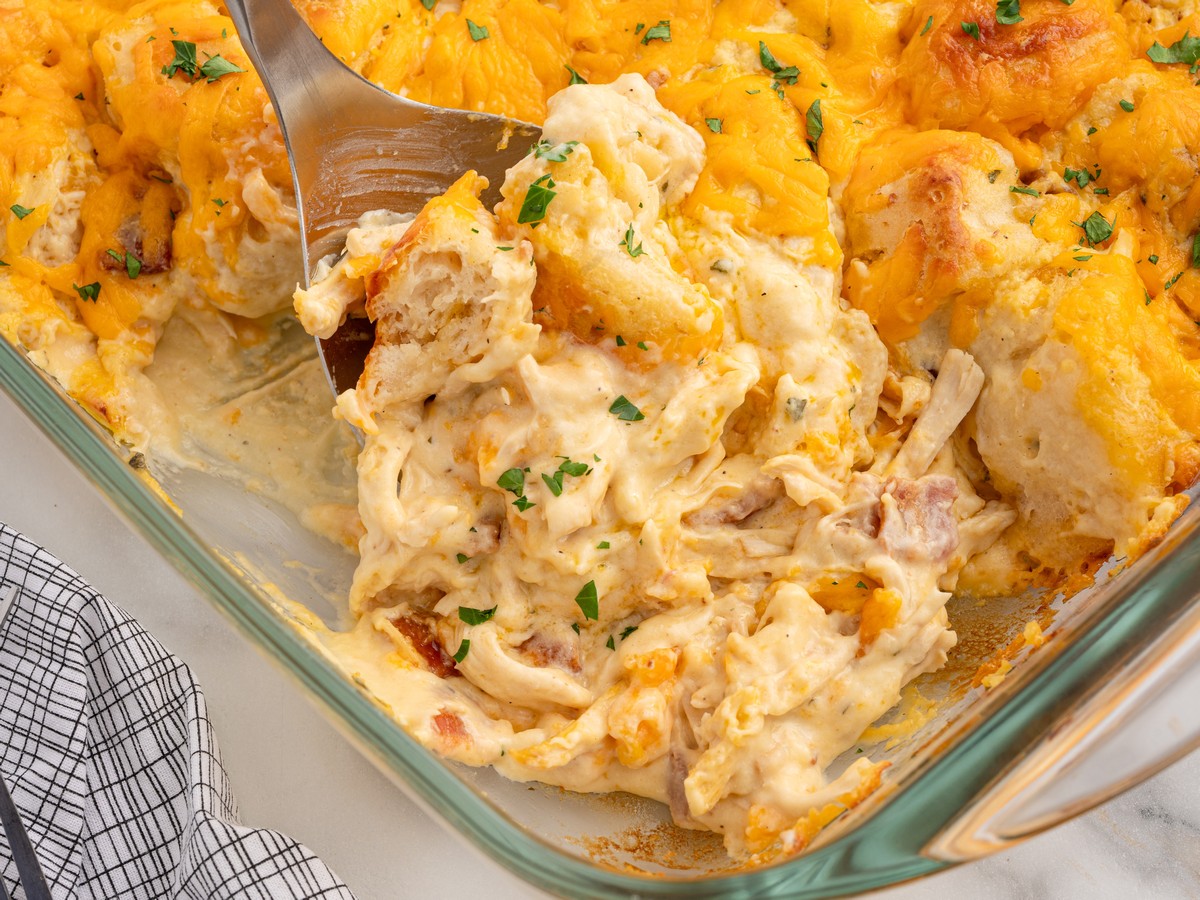 Close-up of Chicken Bubble Biscuit Bake being served from a glass baking dish, showing creamy shredded chicken, bacon, biscuit pieces, melted cheddar cheese, and parsley.