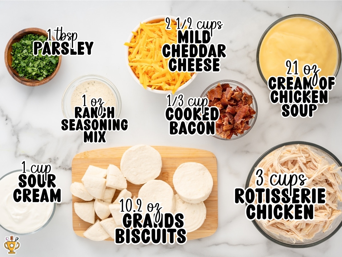 Overhead ingredient display for Chicken Bubble Biscuit Bake labeled 1 Tbsp parsley, 2 ½ cups mild cheddar cheese, 21 oz cream of chicken soup, 1 oz ranch seasoning mix, ⅓ cup cooked bacon, 1 cup sour cream, 10.2 oz Grands biscuits, and 3 cups rotisserie chicken.