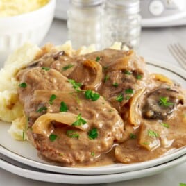 Close-up of Crock Pot Cube Steak with fork-tender beef, mushrooms, onions, and savory gravy served beside mashed potatoes and garnished with parsley.