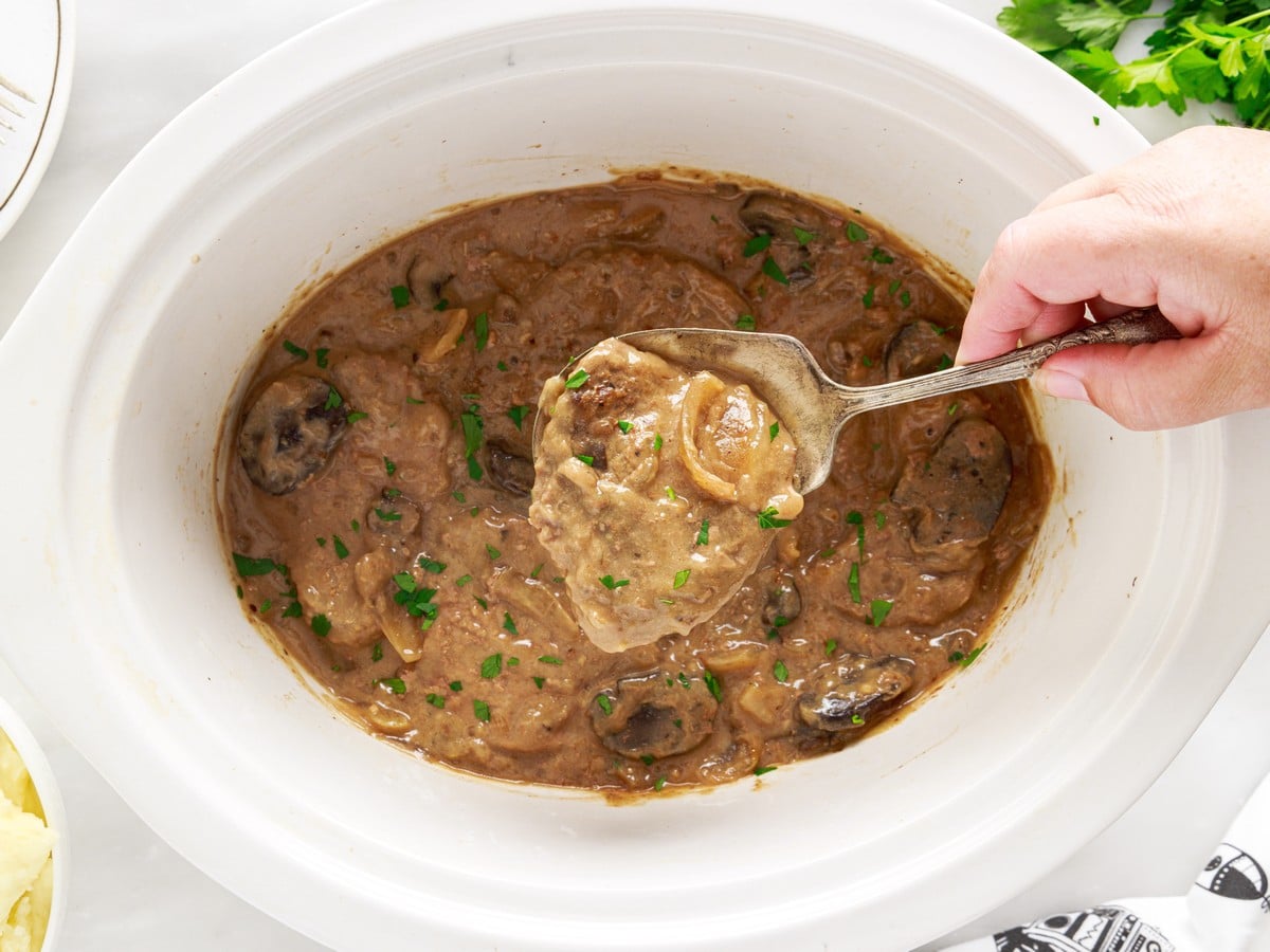 Overhead view of Crock Pot Cube Steak in a white slow cooker, with a spoon lifting tender beef covered in mushroom and onion gravy.