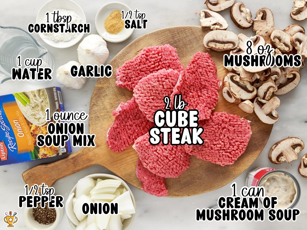 Overhead display of Crock Pot Cube Steak ingredients labeled 1 tablespoon cornstarch, ½ teaspoon salt, 1 cup water, garlic, 8 ounces mushrooms, 1 ounce onion soup mix, 2 pounds cube steak, ½ teaspoon pepper, onion, and 1 can cream of mushroom soup.