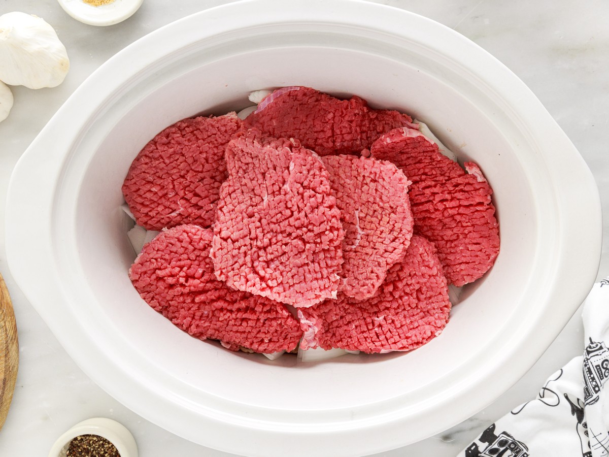 Overhead view of raw cube steaks layered over sliced onions and mushrooms inside a white slow cooker before cooking.