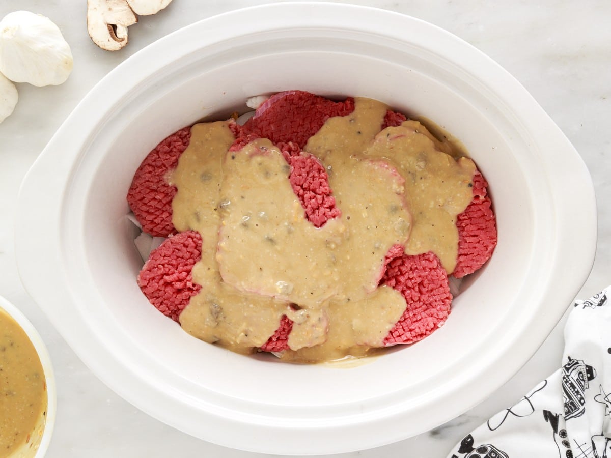 Overhead view of seasoned mushroom gravy poured over raw cube steaks layered in a white slow cooker with onions and mushrooms underneath.