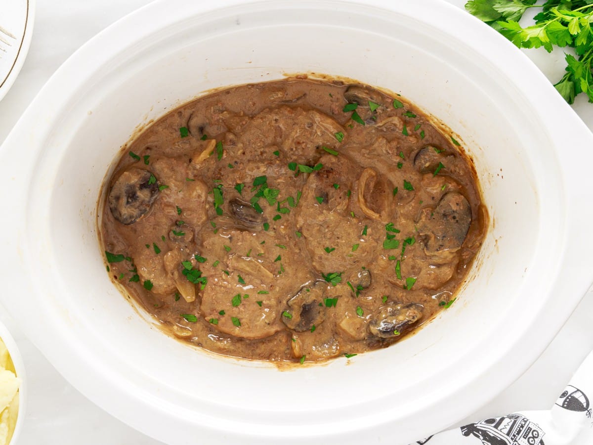 Overhead view of finished Crock Pot Cube Steak in a white slow cooker with tender beef, sliced mushrooms, onions, thick brown gravy, and chopped parsley.