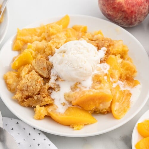 Close-up of Crockpot Peach Dump Cake with juicy peach slices, buttery golden cake topping, and a scoop of melting vanilla ice cream served on a white plate, perfect for an easy slow cooker dessert.