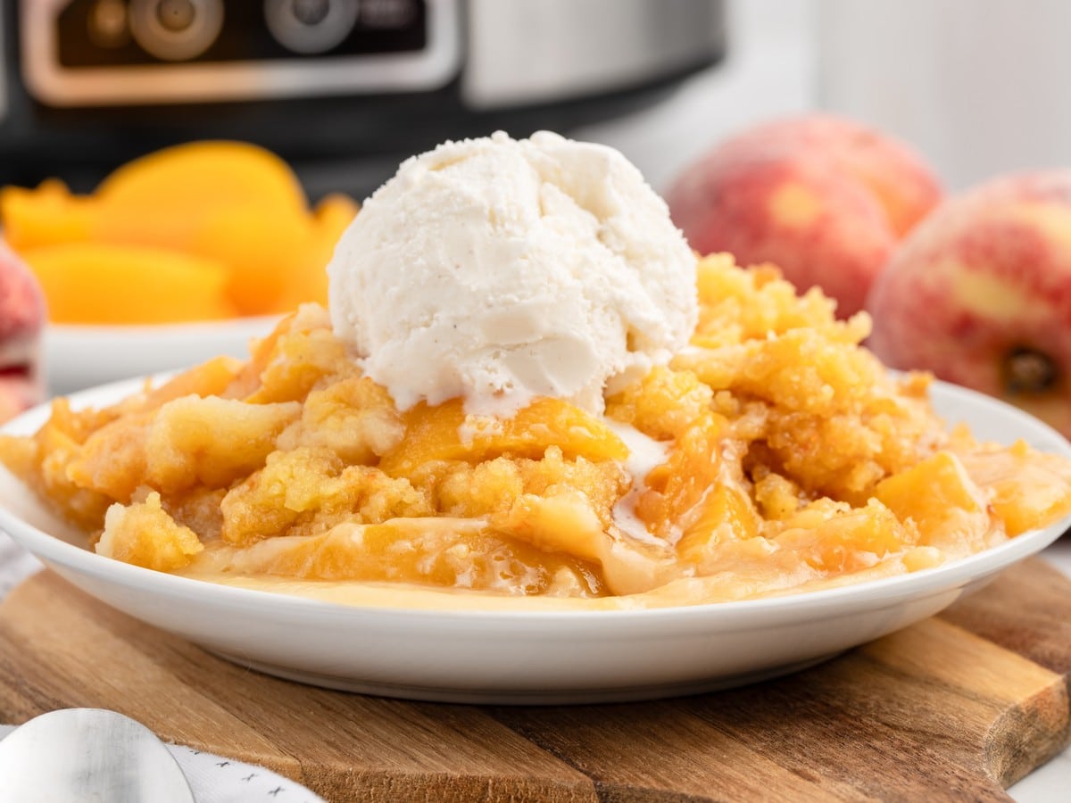 Warm Crockpot Peach Dump Cake topped with a generous scoop of vanilla ice cream on a white plate, with tender peaches, buttery cake topping, and a slow cooker in the background.