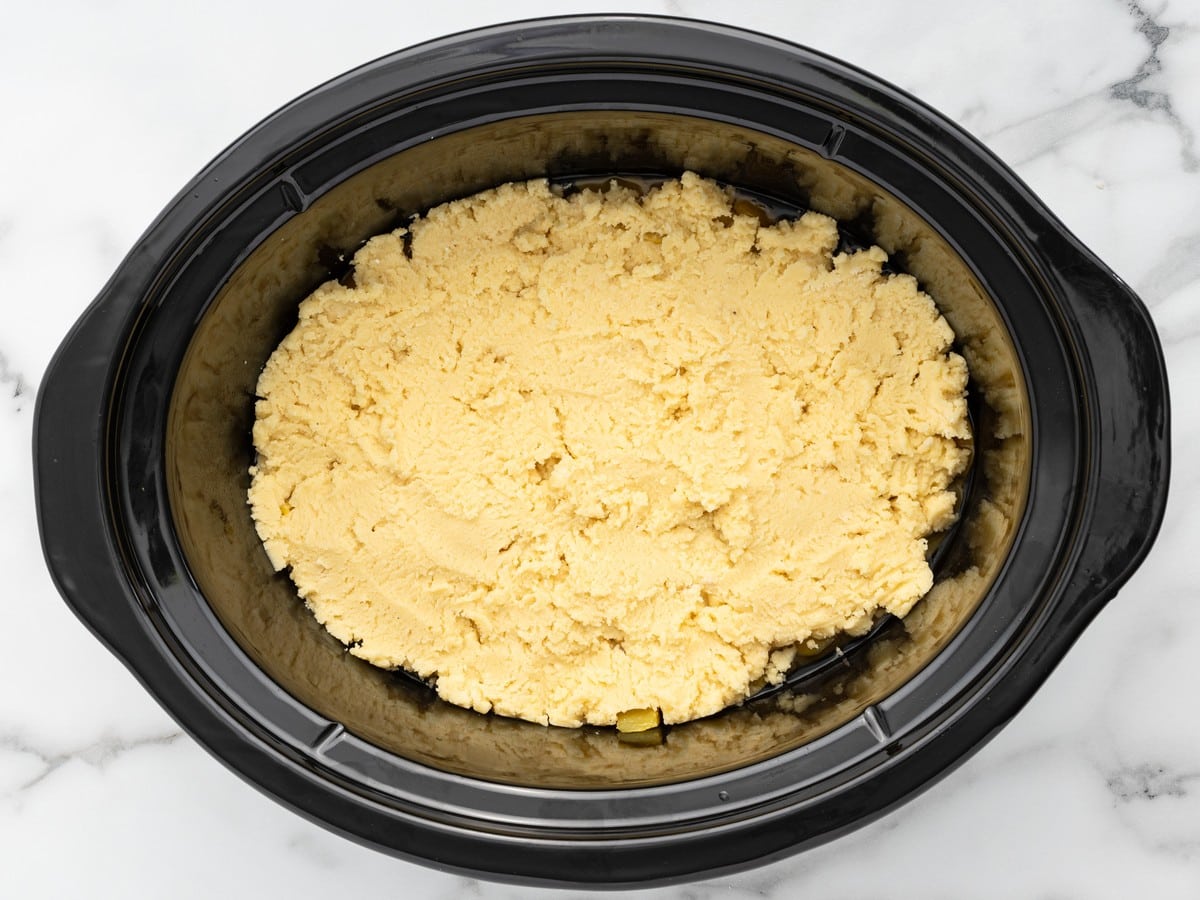 Overhead view of Crockpot Peach Dump Cake before cooking, with the peaches completely covered by an even layer of dry yellow cake mix inside a black slow cooker.