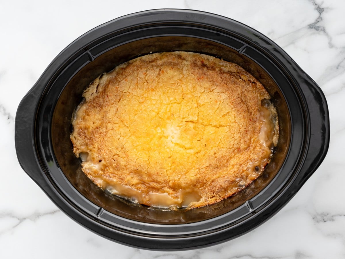 Overhead view of fully cooked Crockpot Peach Dump Cake with a golden buttery cake topping and bubbling peach filling inside a black slow cooker.