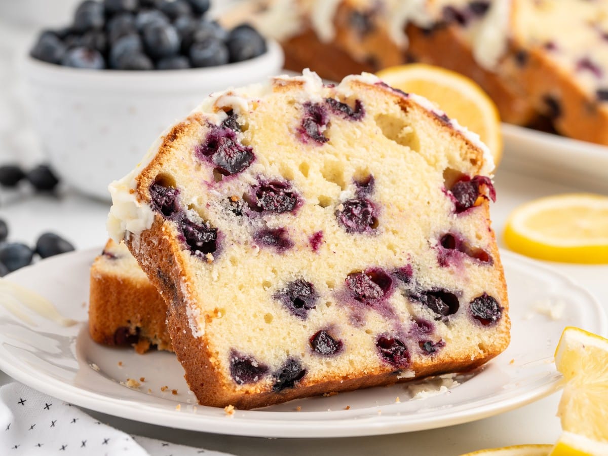 Thick slice of Lemon Blueberry Pound Cake with homemade lemon glaze, showcasing a tender buttery crumb filled with juicy blueberries, served on a white plate with fresh lemon wedges.