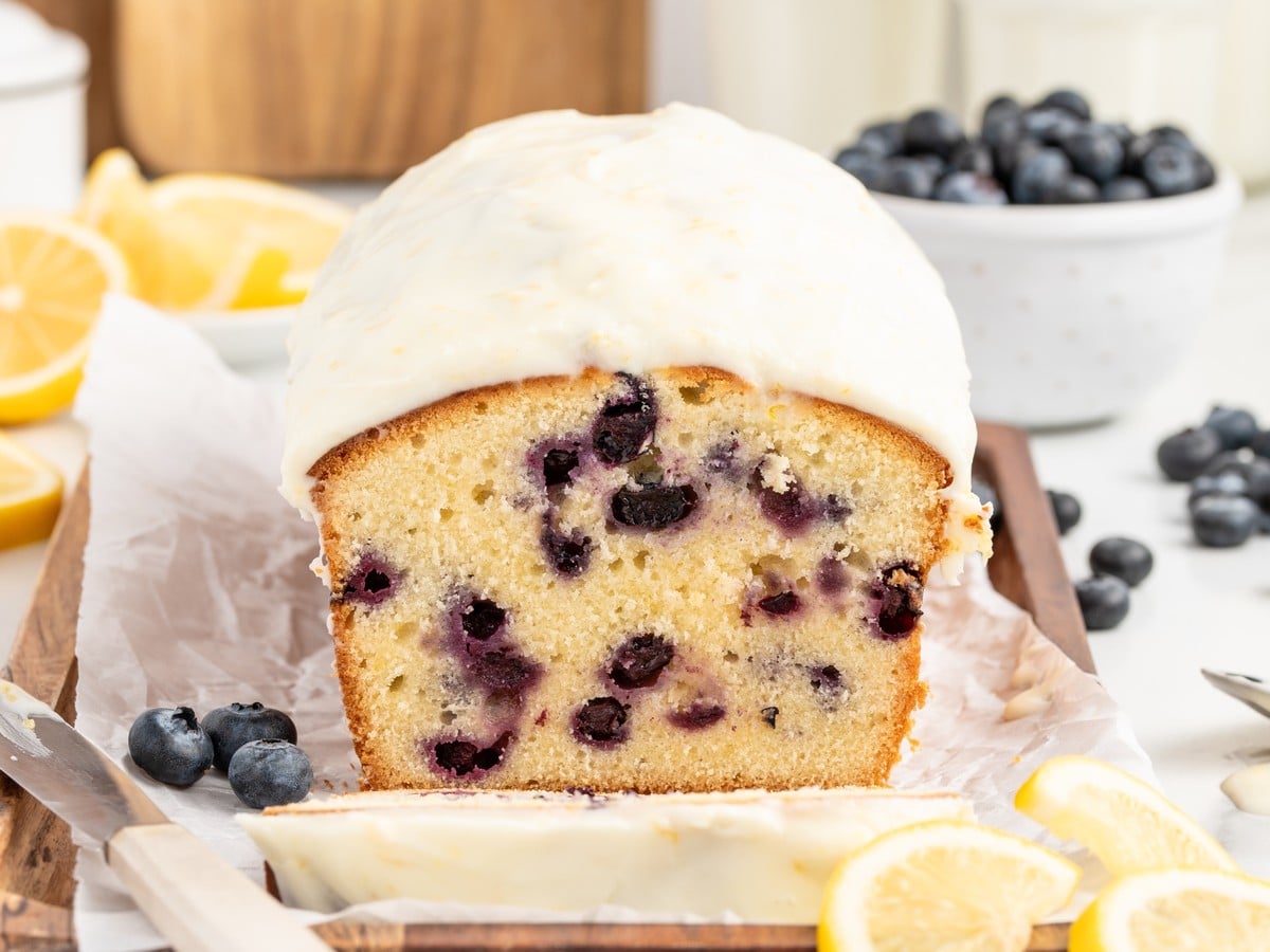 Front view of a whole Lemon Blueberry Pound Cake topped with a thick homemade lemon glaze, revealing a moist, buttery crumb packed with fresh blueberries and garnished with lemon slices.