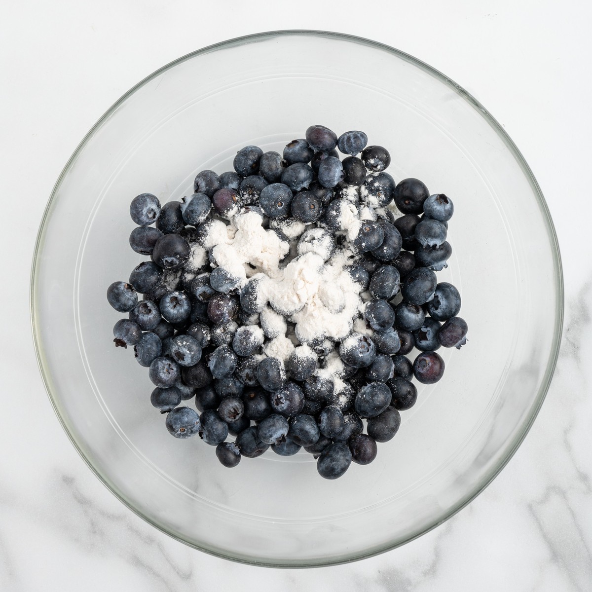 Fresh blueberries tossed with flour in a clear glass mixing bowl to coat them before folding into Lemon Blueberry Pound Cake batter, helping keep the berries evenly suspended while baking.