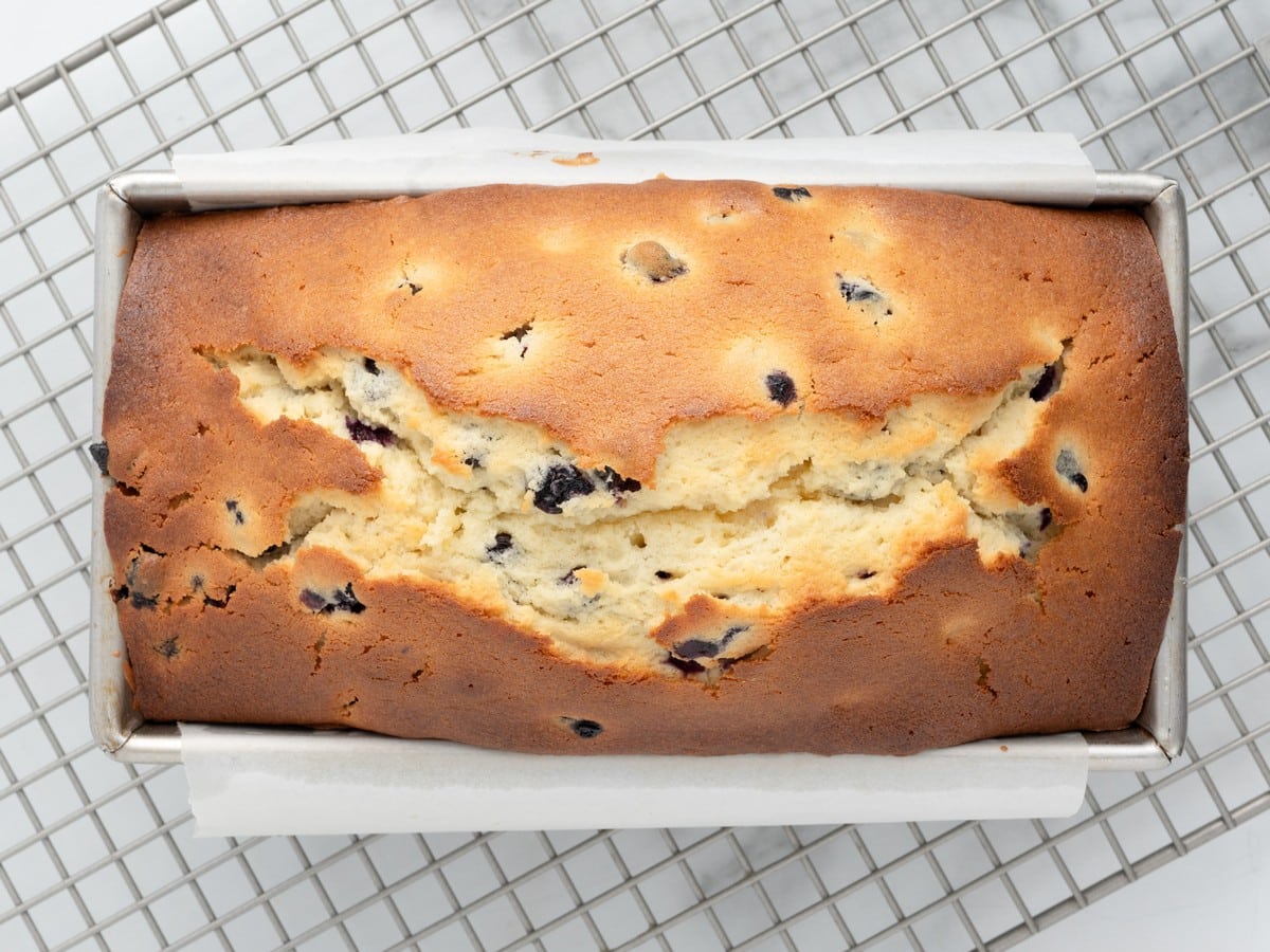 Freshly baked Lemon Blueberry Pound Cake cooling in a loaf pan on a wire rack, showing a golden brown crust with the signature crack across the top and blueberries peeking through.