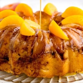 Close-up of Peach Cobbler Pound Cake topped with juicy peach slices while a glossy peach glaze is poured over the golden cinnamon-swirled Bundt cake.