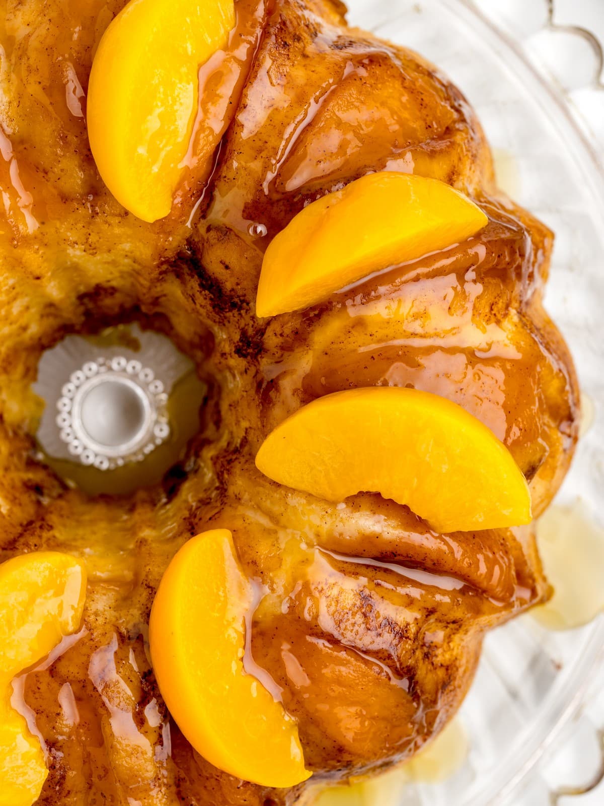 Close-up overhead view of Peach Cobbler Pound Cake topped with bright peach slices, cinnamon, and glossy caramel sauce on a glass cake stand.