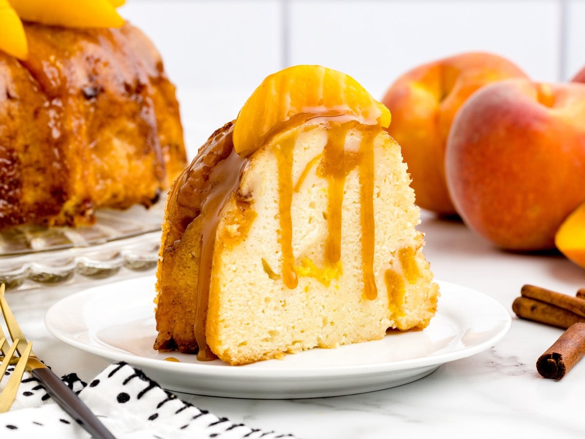 Slice of Peach Cobbler Pound Cake on a white plate, showing a dense buttery crumb with peach pieces, caramel drizzle, and a peach slice on top.