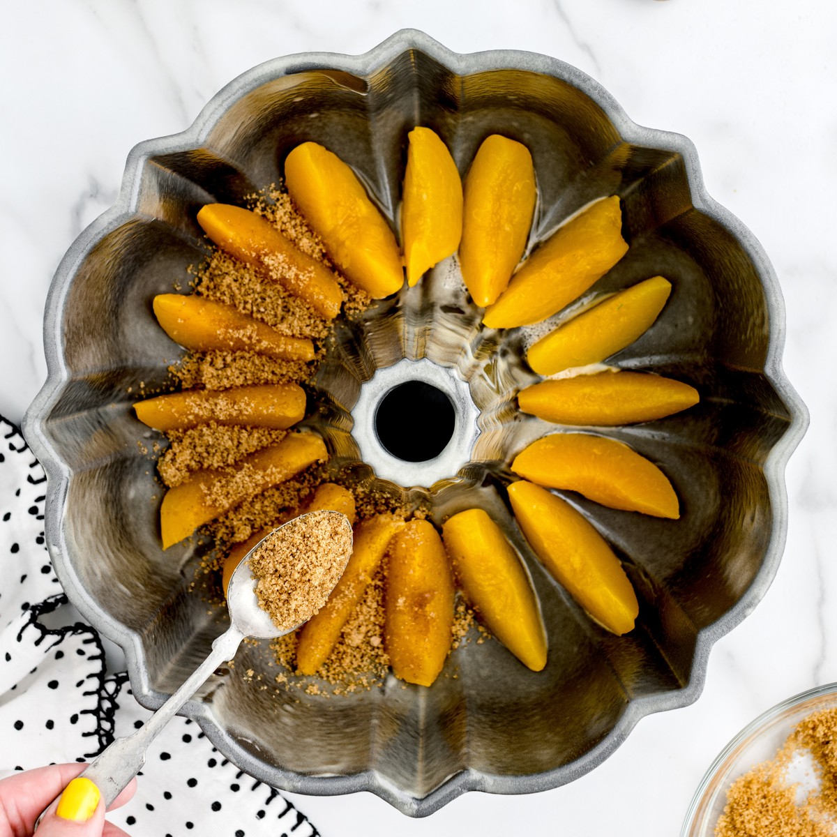 Overhead view of peach slices arranged around the bottom of a greased Bundt pan while brown sugar is sprinkled over them for Peach Cobbler Pound Cake.