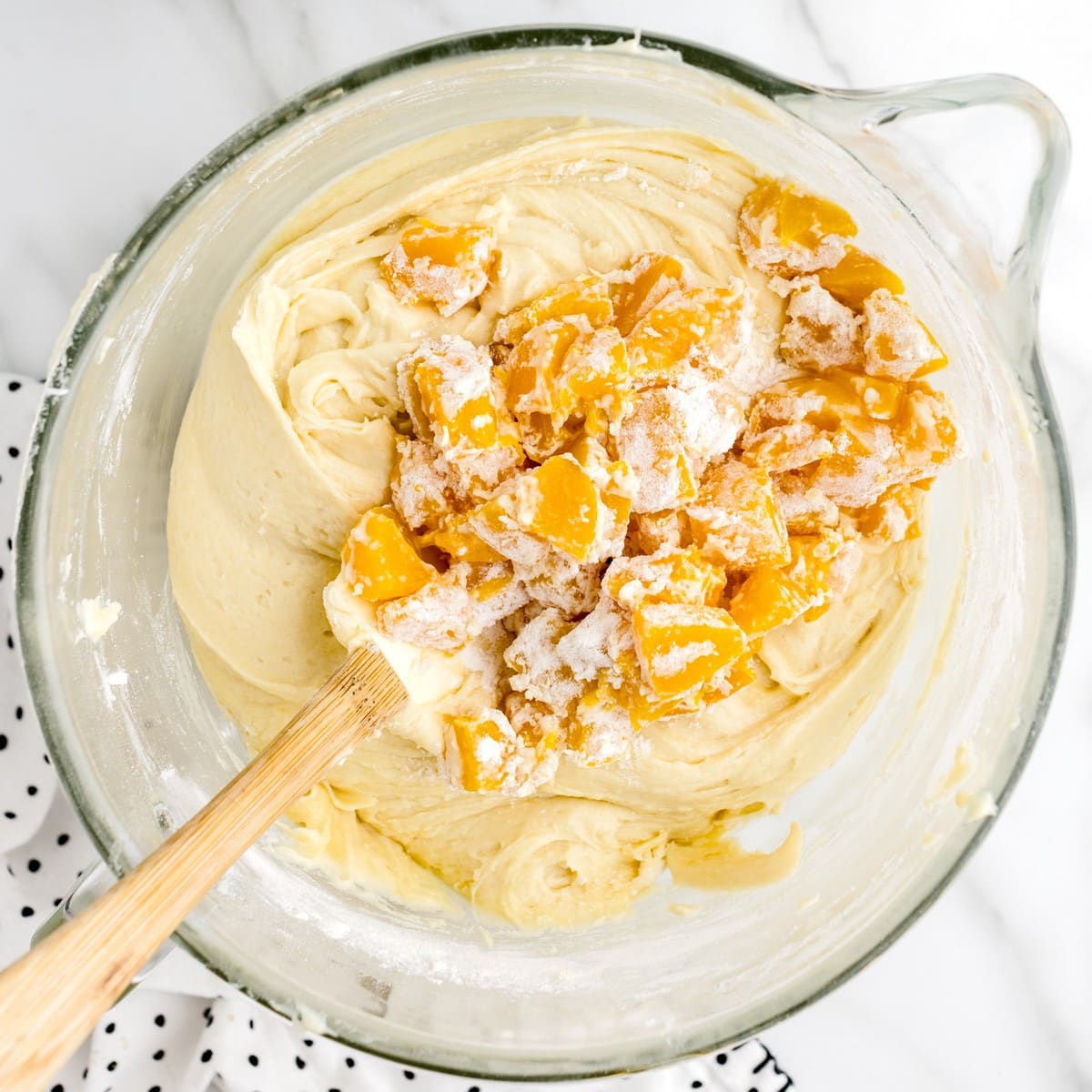 Overhead view of flour-coated peach pieces being folded into thick pound cake batter in a glass mixing bowl for Peach Cobbler Pound Cake.