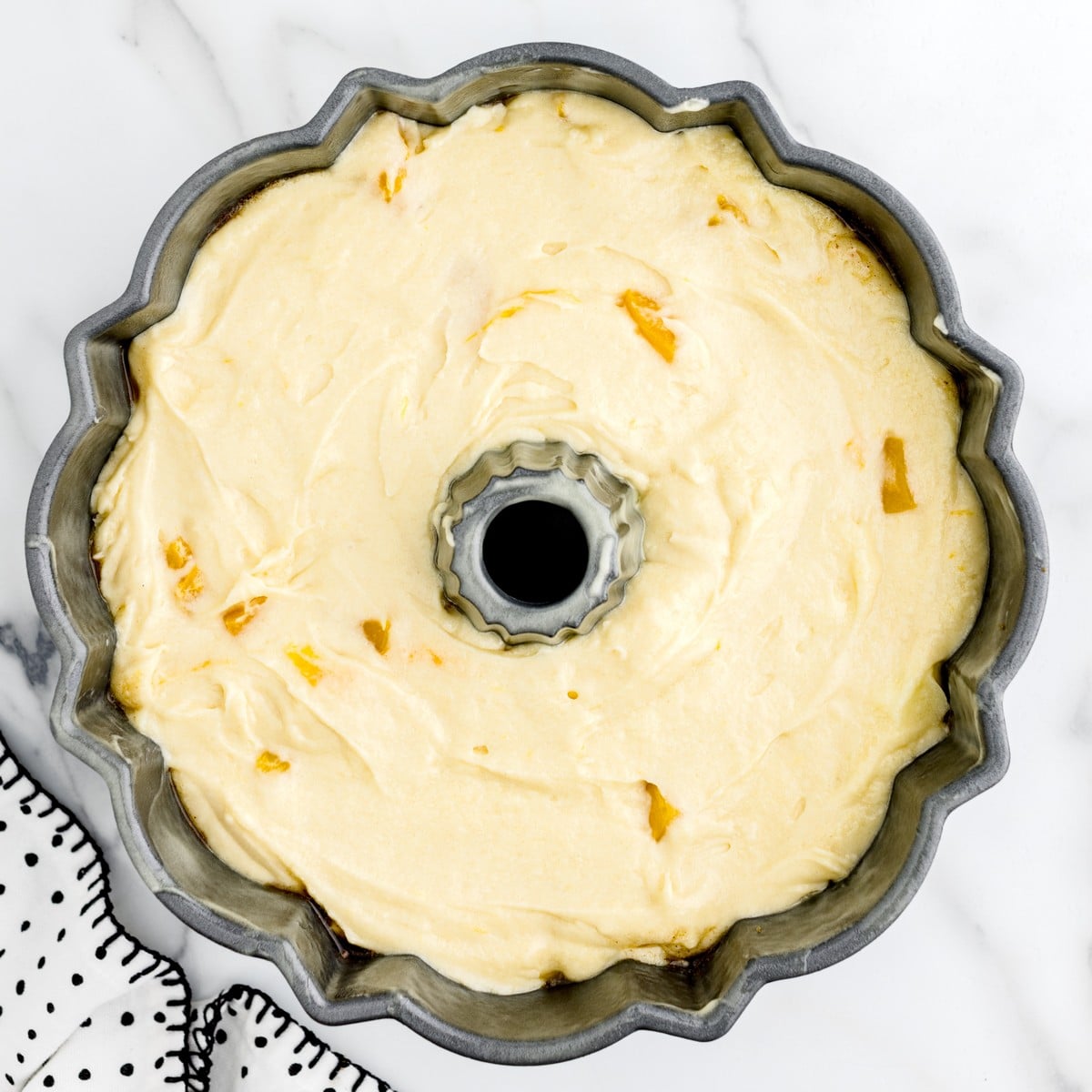 Overhead view of Peach Cobbler Pound Cake batter spread evenly in a prepared Bundt pan with small peach pieces visible throughout.