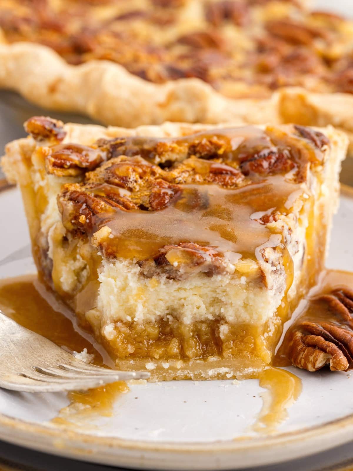 Close-up of a thick slice of Pecan Cheesecake Pie showing a flaky crust, gooey pecan filling, creamy cheesecake center, toasted pecans, and caramel sauce drizzled over the top.