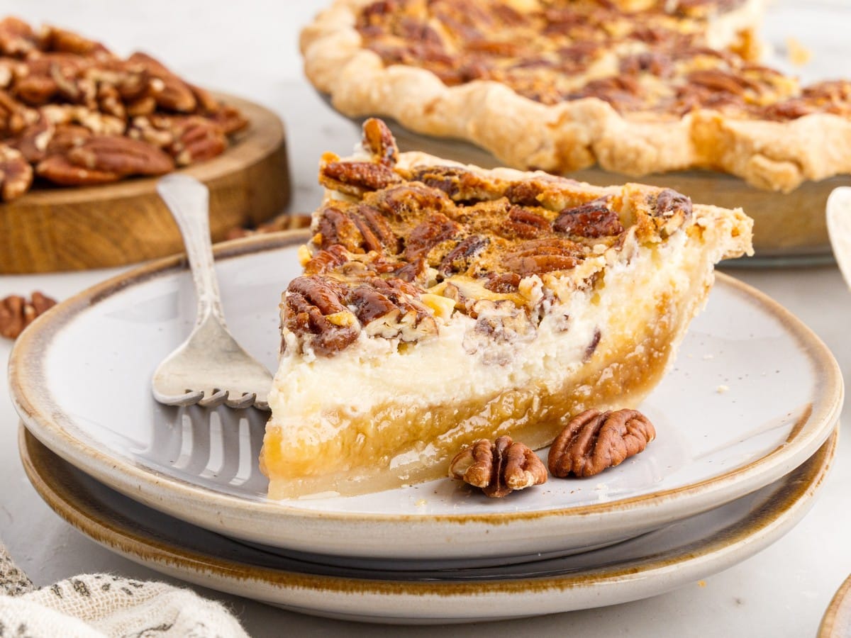 Side view of a thick slice of Pecan Cheesecake Pie on a ceramic plate, showing a flaky crust, gooey pecan pie layer, creamy cheesecake filling, and toasted pecans on top.