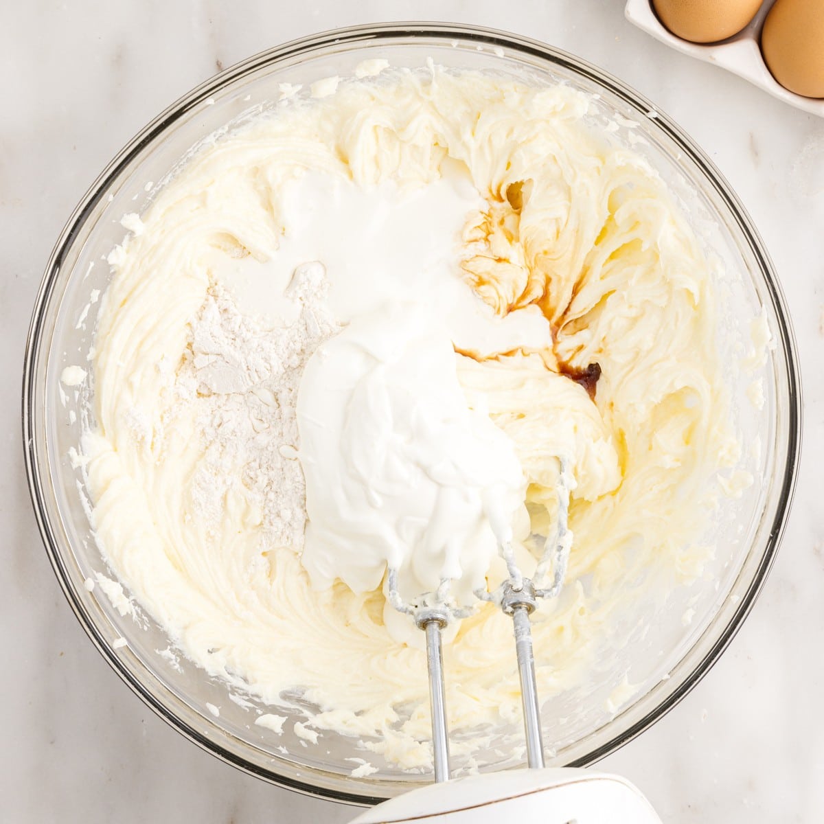 Overhead view of flour, sour cream, heavy whipping cream, and vanilla added to whipped cream cheese in a glass mixing bowl while preparing the cheesecake layer for Pecan Cheesecake Pie.
