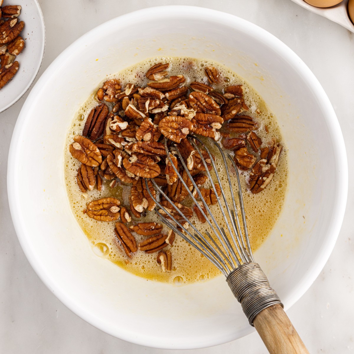 Overhead view of pecans being stirred into the sweet egg and corn syrup filling for Pecan Cheesecake Pie, with a metal whisk resting inside a white mixing bowl.