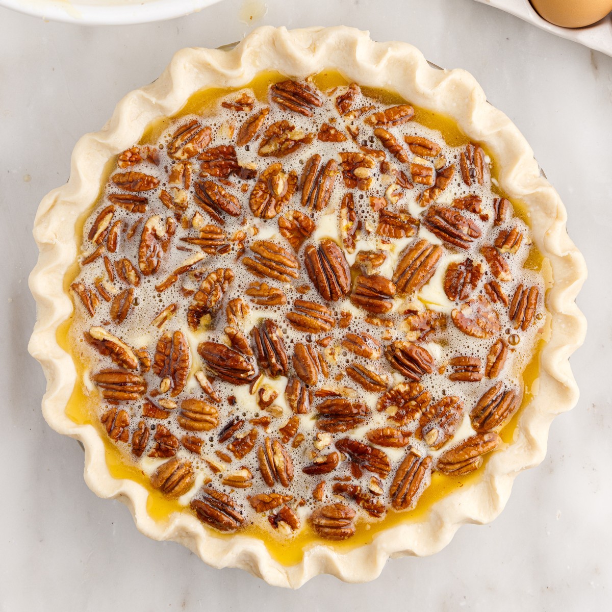 Overhead view of an unbaked Pecan Cheesecake Pie filled with creamy batter and pecan pie mixture, topped with whole and chopped pecans inside a fluted pie crust.