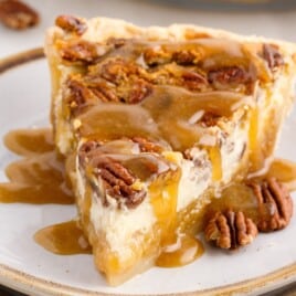 A slice of Pecan Cheesecake Pie served on a ceramic plate with creamy cheesecake filling, a gooey pecan layer, flaky crust, whole pecans, and caramel sauce drizzled over the top.