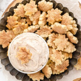 Overhead view of cinnamon-sugar pie crust chips shaped like autumn leaves, arranged around a bowl of creamy cinnamon dip on a dark serving platter.