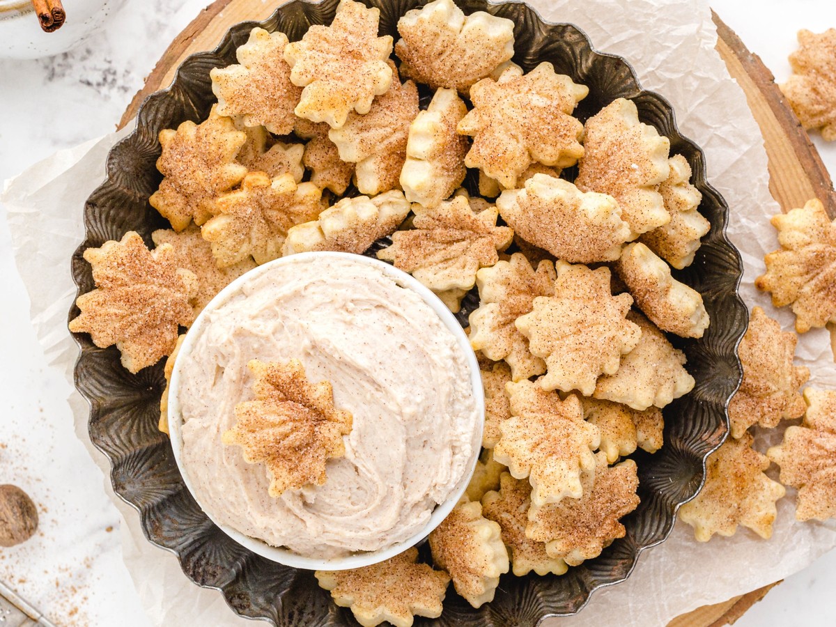 Overhead view of cinnamon-sugar pie crust chips shaped like autumn leaves, arranged around a bowl of creamy cinnamon dip on a dark serving platter.