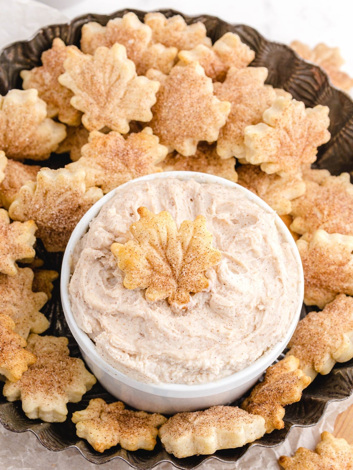 Overhead view of leaf-shaped cinnamon-sugar pie crust chips arranged around a bowl of creamy cinnamon dip on a dark decorative platter.