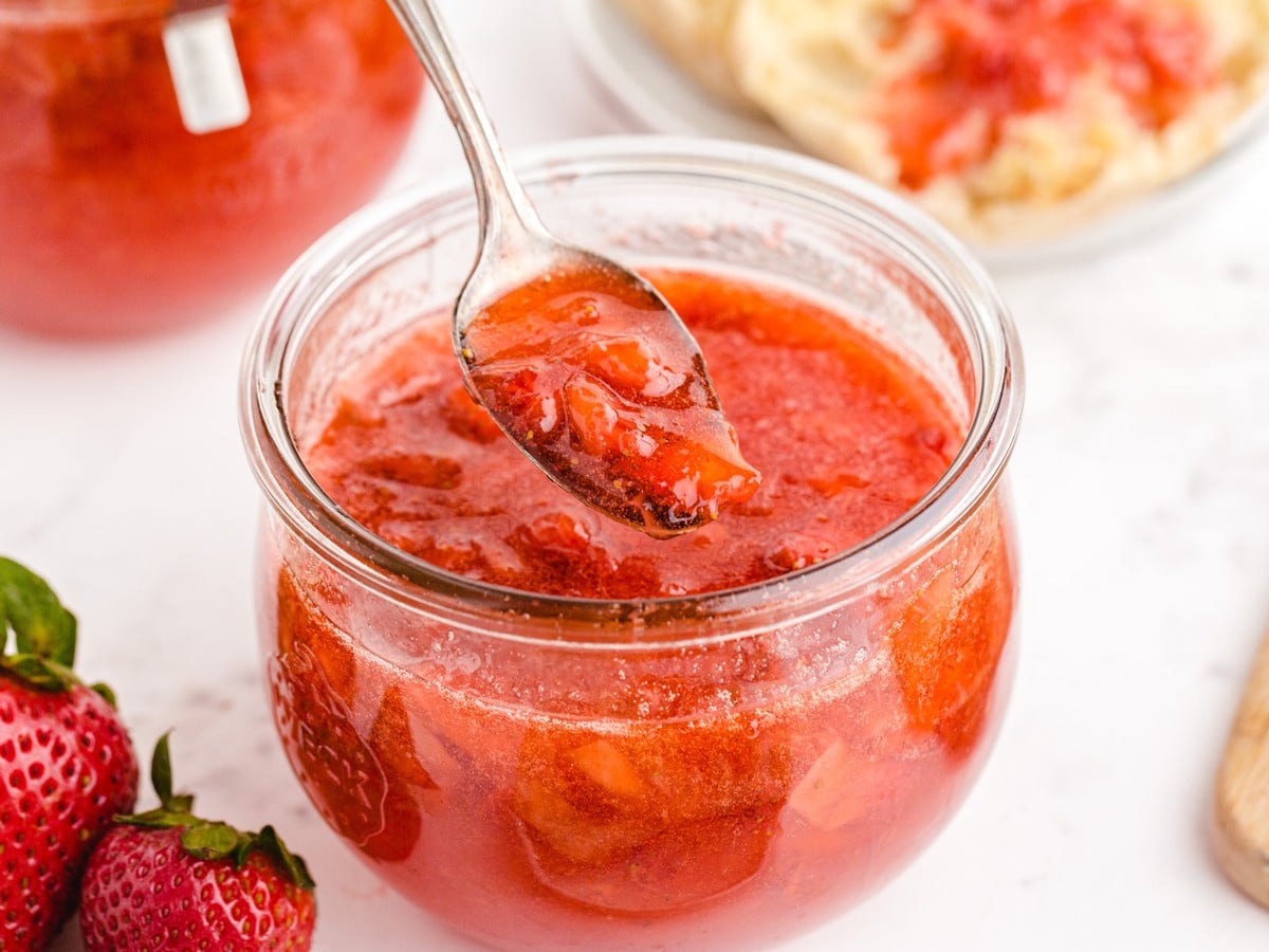 Homemade Strawberry Freezer Jam in a glass jar with a spoon lifting chunky strawberry preserves, served with fresh strawberries and toast in the background.