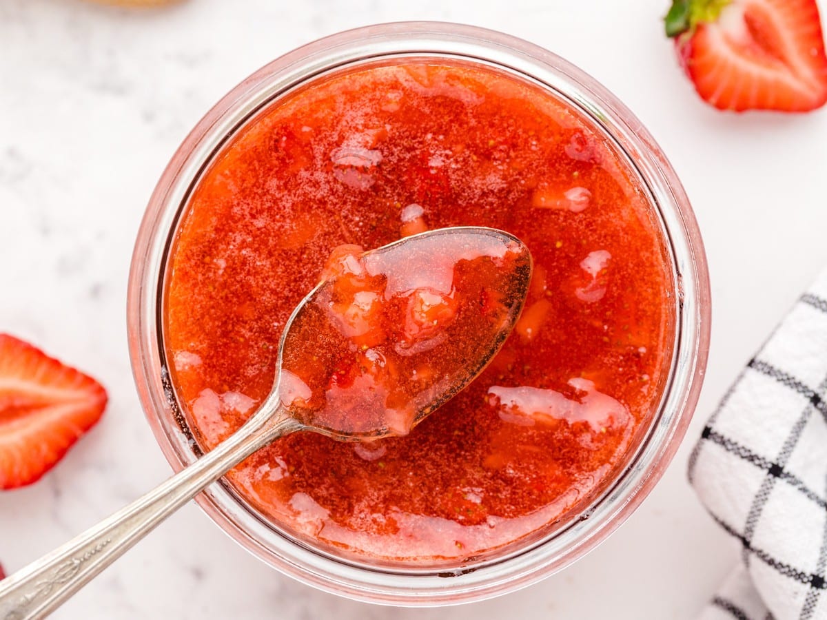 Overhead view of homemade Strawberry Freezer Jam in a glass jar with a spoon, showing a bright red, chunky strawberry texture.