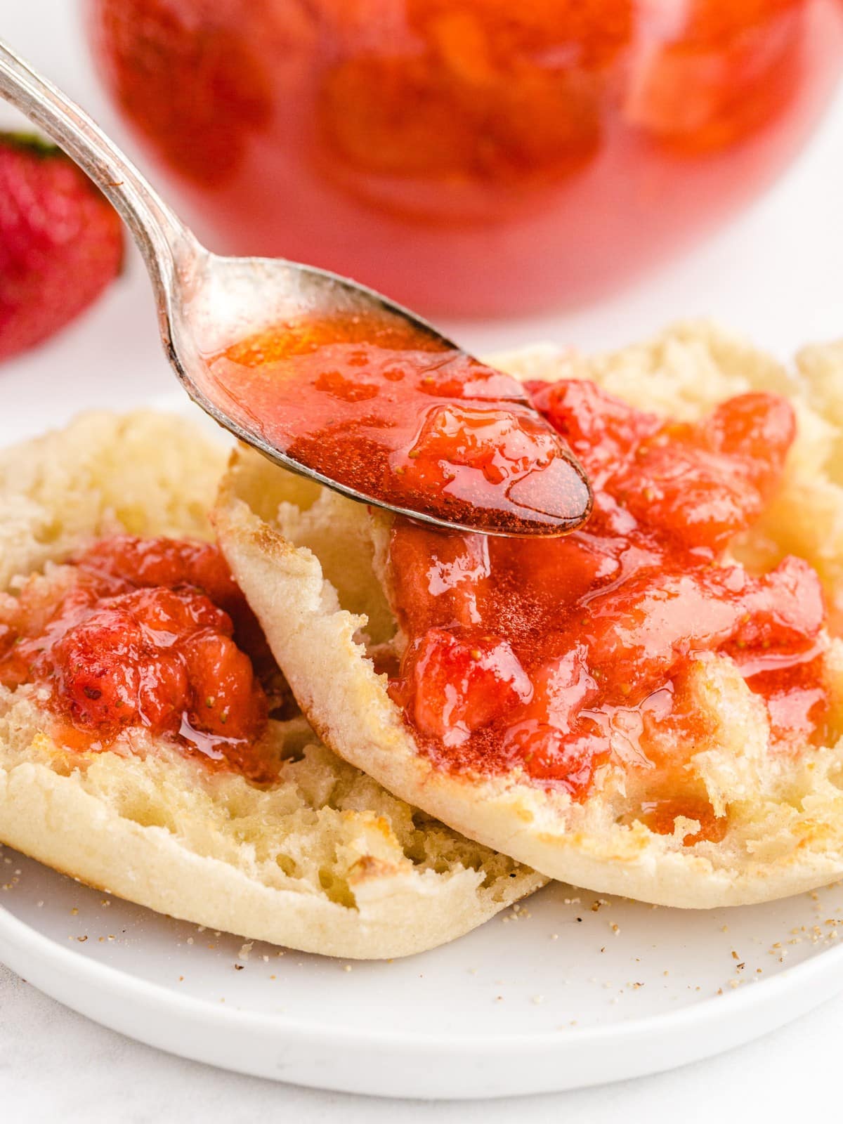 Spoon spreading homemade Strawberry Freezer Jam with juicy strawberry pieces onto a warm English muffin for an easy breakfast or snack.