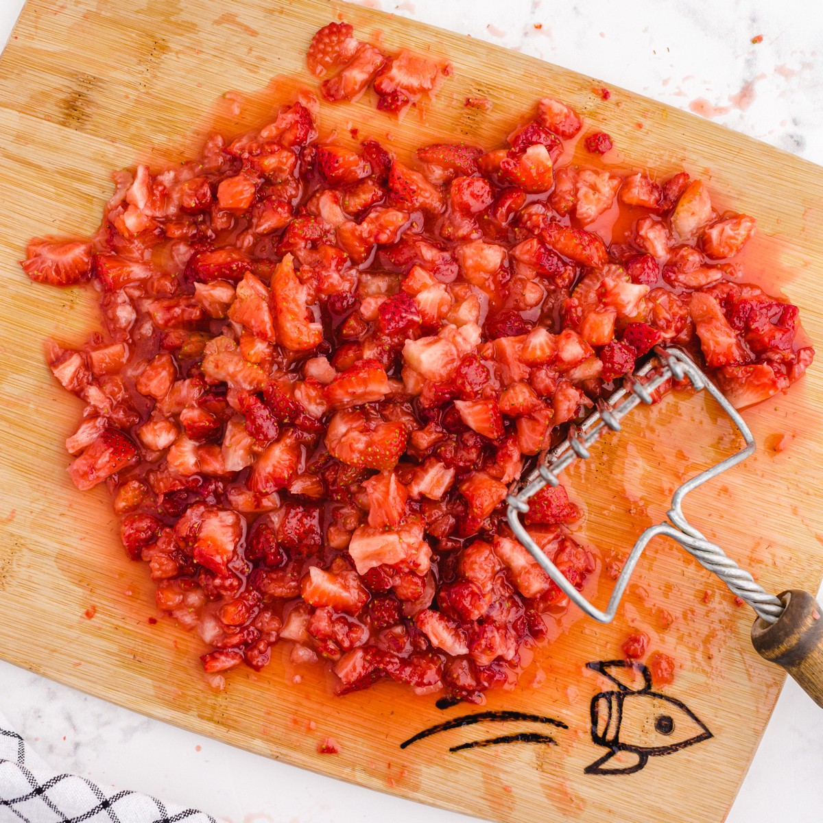Fresh strawberries crushed with a potato masher on a cutting board, creating small juicy pieces for homemade Strawberry Freezer Jam.