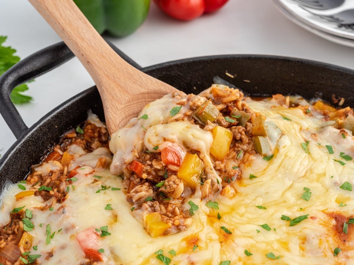 Close-up of a wooden spoon lifting a cheesy scoop of Stuffed Pepper Casserole with ground beef, rice, diced bell peppers, and melted mozzarella from a cast iron skillet.
