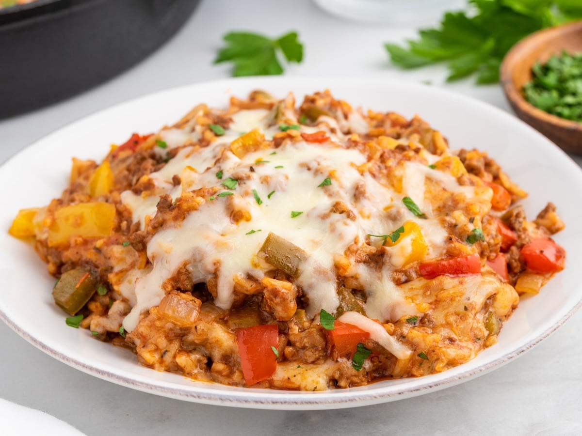 Side view of Stuffed Pepper Casserole served on a white plate with melted mozzarella, seasoned ground beef, rice, colorful bell peppers, onions, and fresh parsley garnish.