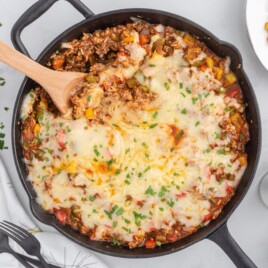 Wooden spoon lifting a cheesy serving of Stuffed Pepper Casserole from a cast iron skillet topped with melted Monterey Jack cheese and fresh parsley.
