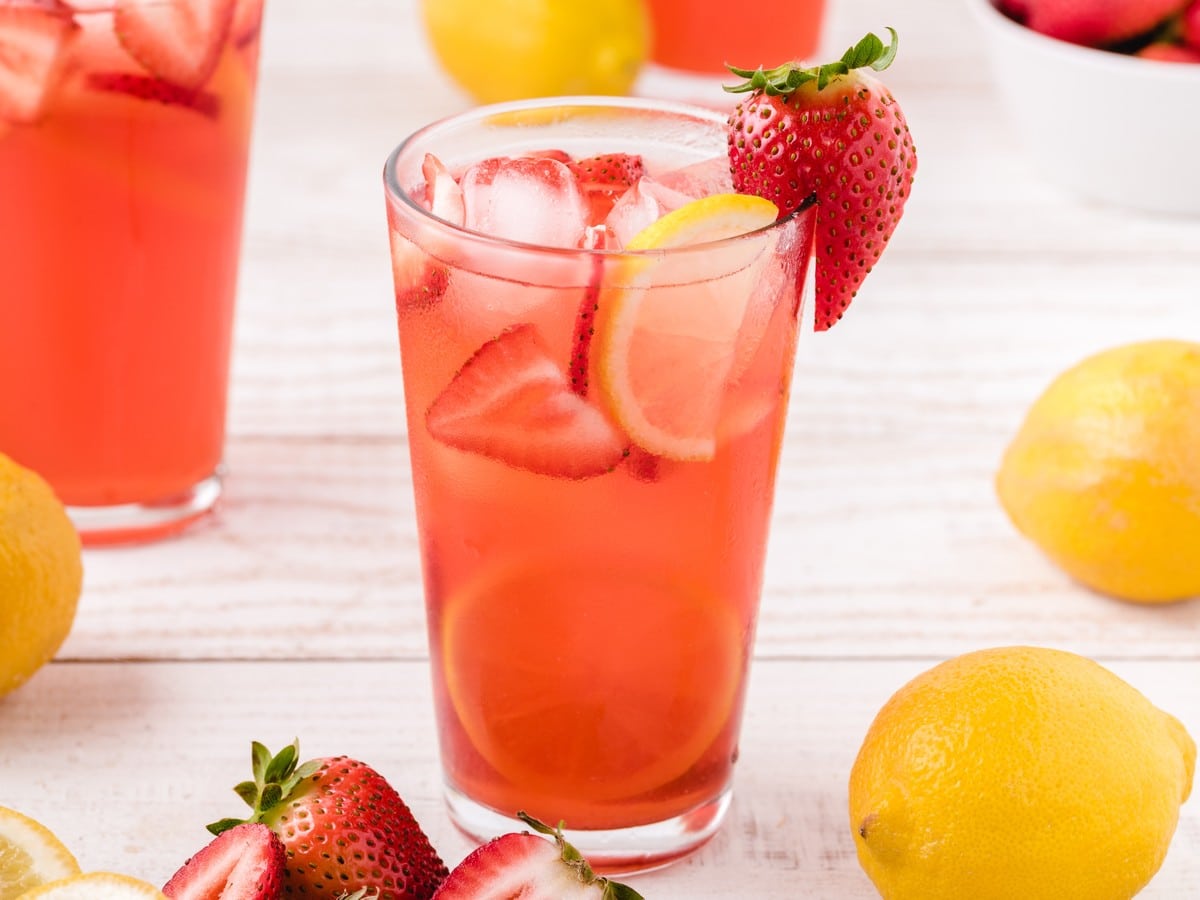 Glass of Strawberry Lemonade with ice, sliced strawberries, lemon slices, and a fresh strawberry garnish on a bright summer drink setup.