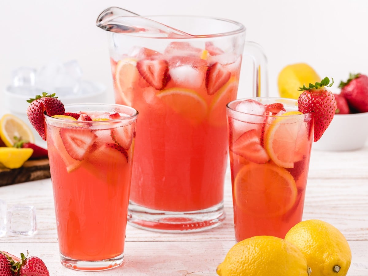 Pitcher and two glasses of Strawberry Lemonade filled with ice, sliced strawberries, fresh lemon slices, and strawberry garnishes on a bright summer table.