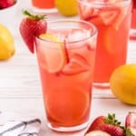 Refreshing homemade Strawberry Lemonade served over ice with fresh strawberry slices and lemon slices in a tall glass, with whole lemons and fresh strawberries in the background.