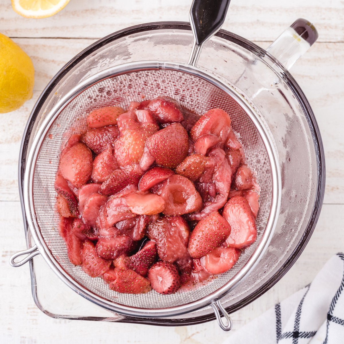 Cooked strawberries being strained through a fine mesh sieve to separate the homemade strawberry syrup for Strawberry Lemonade.