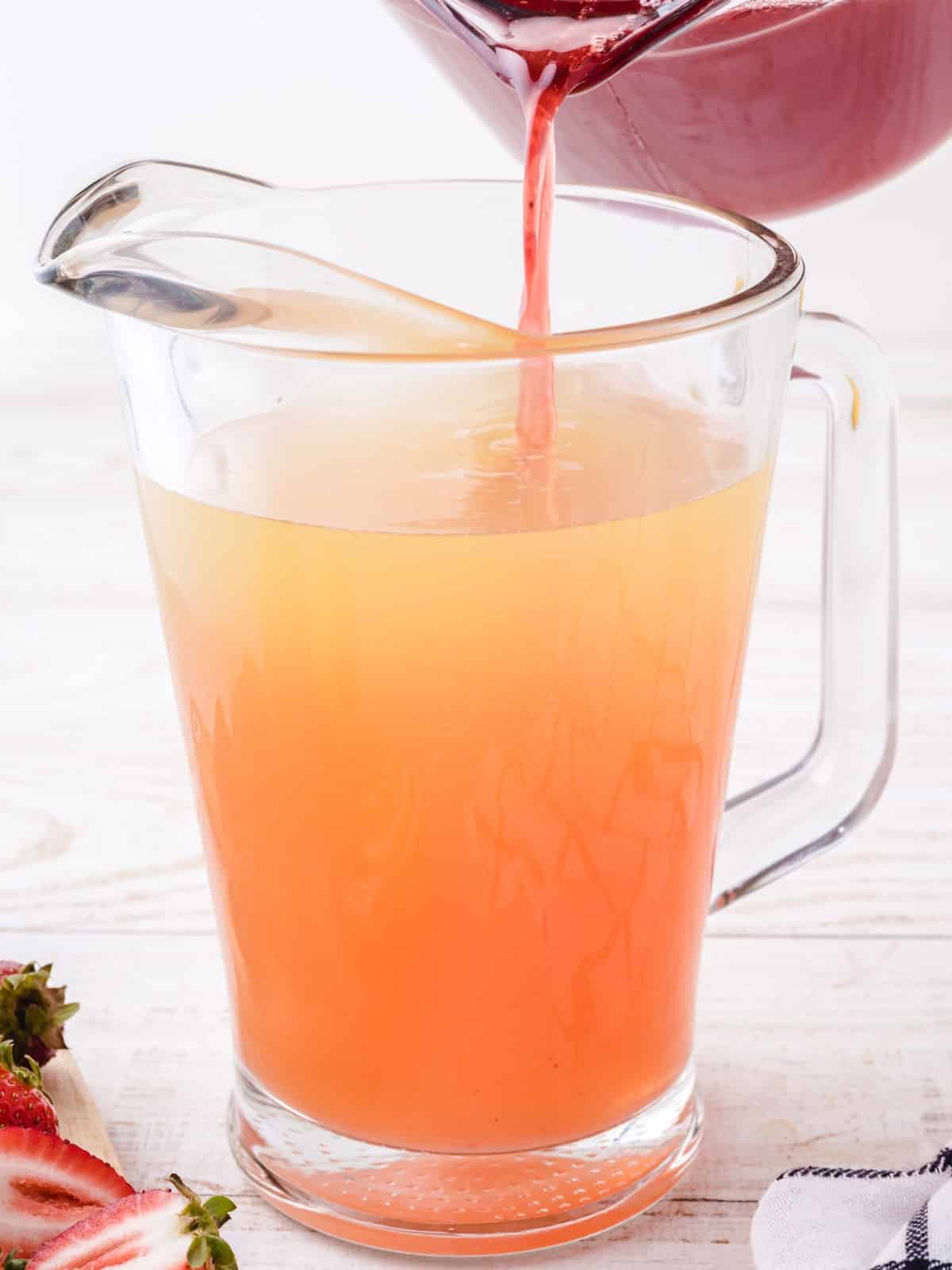 Pouring homemade strawberry syrup into a pitcher of prepared lemonade to make fresh Strawberry Lemonade.