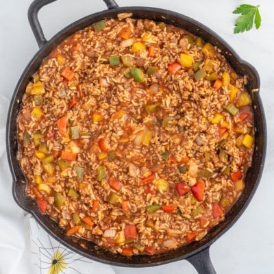 Overhead shot of stuffed pepper casserole in a cast iron skillet, showing ground beef, jasmine rice, and diced red, yellow, and green bell peppers in a rich tomato sauce, garnished with a sprig of fresh parsley on the side.