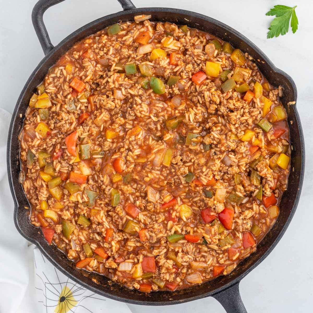 Overhead shot of stuffed pepper casserole in a cast iron skillet, showing ground beef, jasmine rice, and diced red, yellow, and green bell peppers in a rich tomato sauce, garnished with a sprig of fresh parsley on the side.