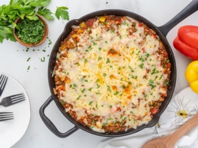 Overhead shot of finished stuffed pepper casserole in a cast iron skillet, topped with melted Monterey Jack cheese and chopped fresh parsley, with diced red, yellow, and green bell peppers visible around the edges, surrounded by fresh parsley, forks on a white plate, and whole red and yellow bell peppers.