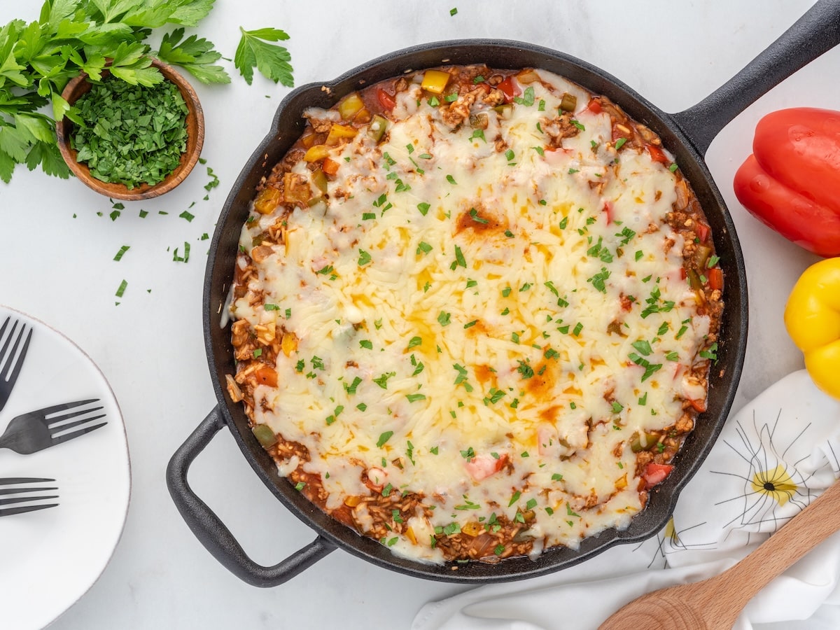 Overhead shot of finished stuffed pepper casserole in a cast iron skillet, topped with melted Monterey Jack cheese and chopped fresh parsley, with diced red, yellow, and green bell peppers visible around the edges, surrounded by fresh parsley, forks on a white plate, and whole red and yellow bell peppers.