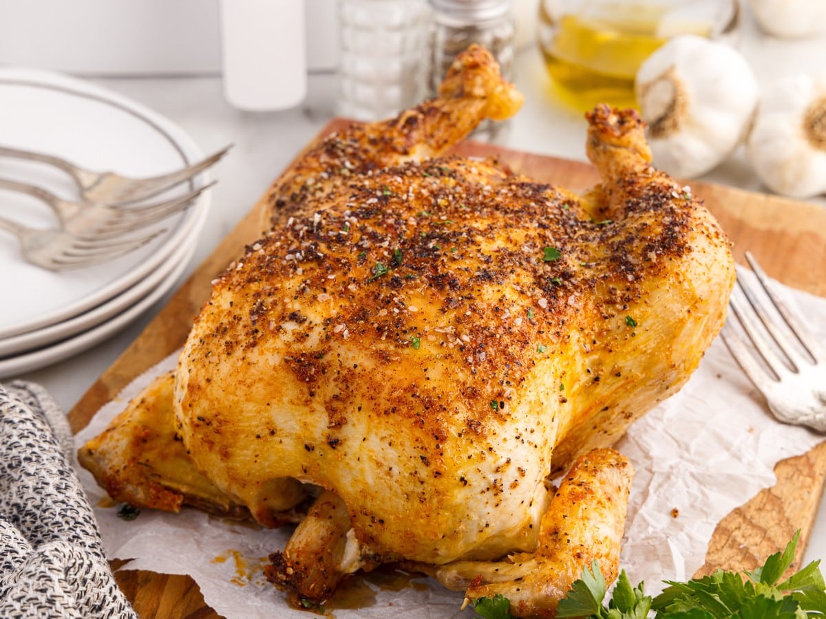 Golden-brown Air Fryer Whole Chicken resting on a wooden cutting board with crisp seasoned skin, fresh parsley, garlic bulbs, and serving forks nearby.