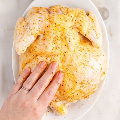 Raw whole chicken being rubbed with olive oil and seasonings by hand on a white platter while preparing Air Fryer Whole Chicken.